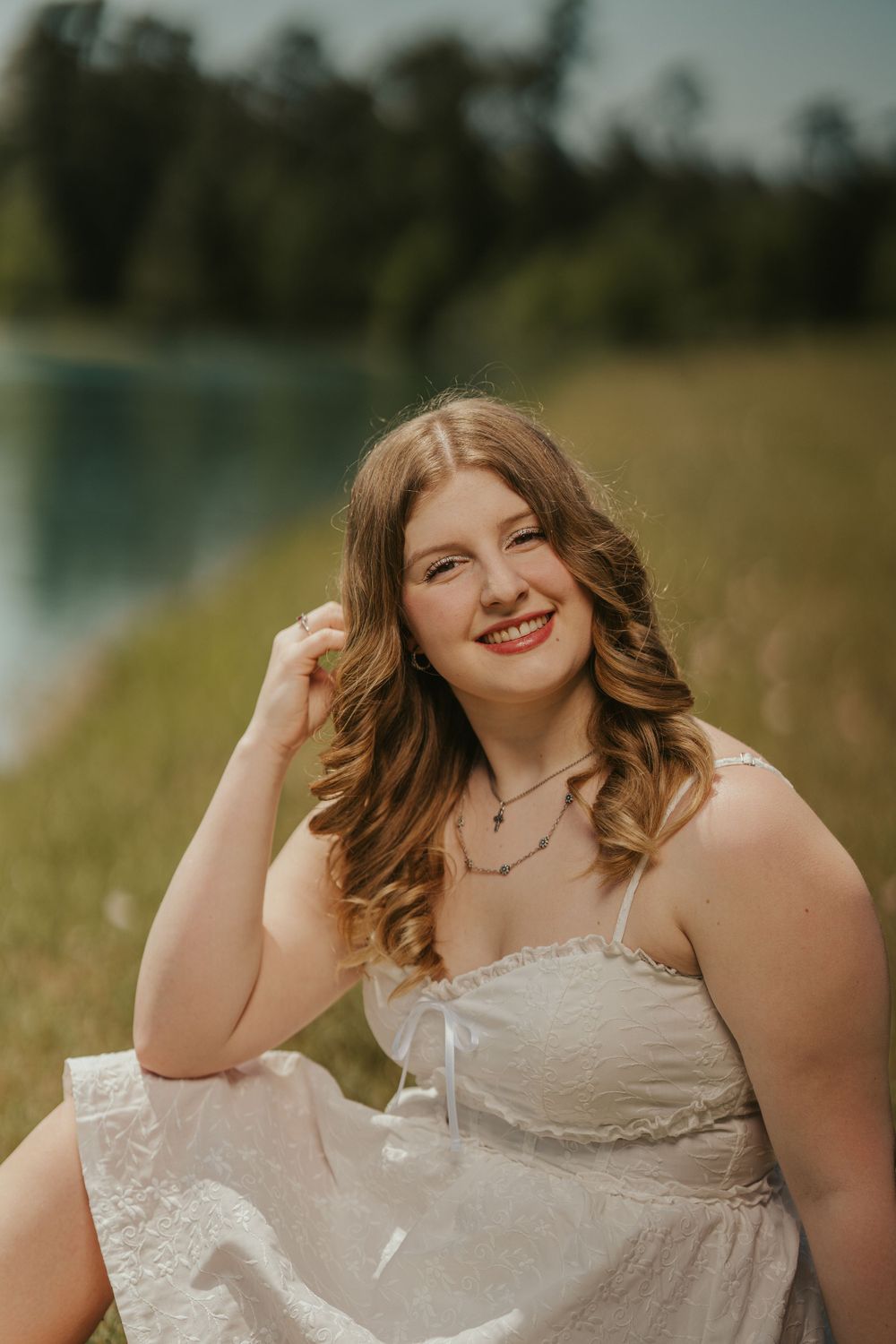 A person in a white strapless dress sits in a grassy field near a lake, with soft sunlight and nature in the background.