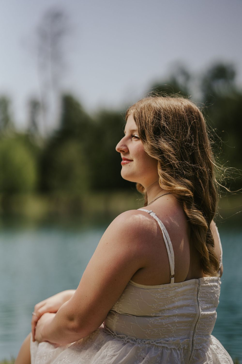 A woman in a pale summer dress sits by a serene lake on a sunny day with trees in the background.