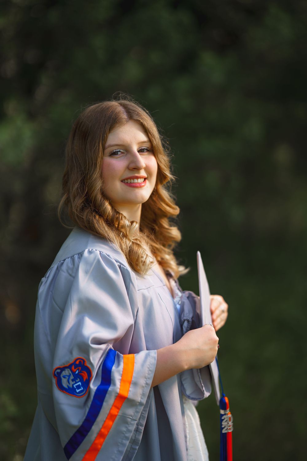 Graduate wearing light gray academic gown with blue and orange honor cords poses outdoors with natural background.