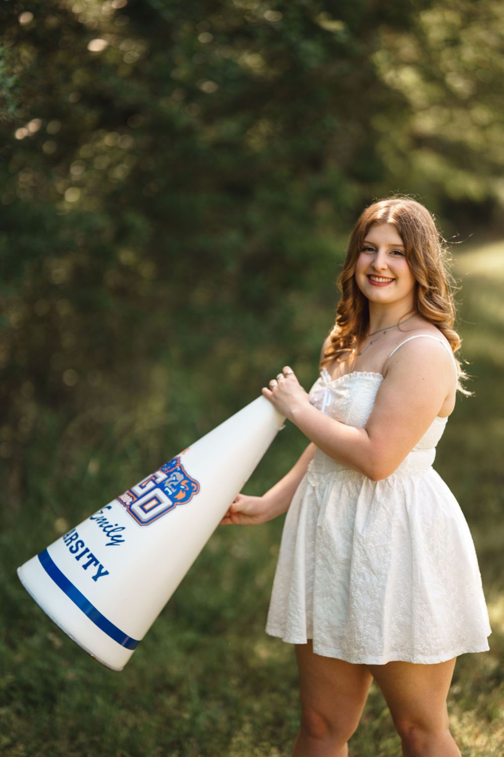 A person in a white summer dress poses outdoors holding a large cheerleading megaphone with blue details.