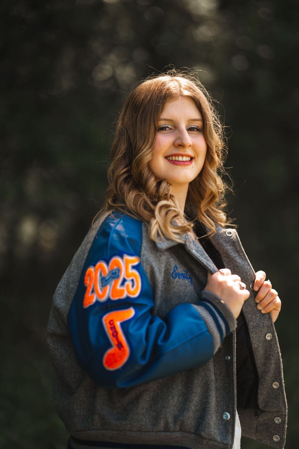 Person wearing a blue and gray letterman jacket with musical note design smiles outdoors in natural lighting.