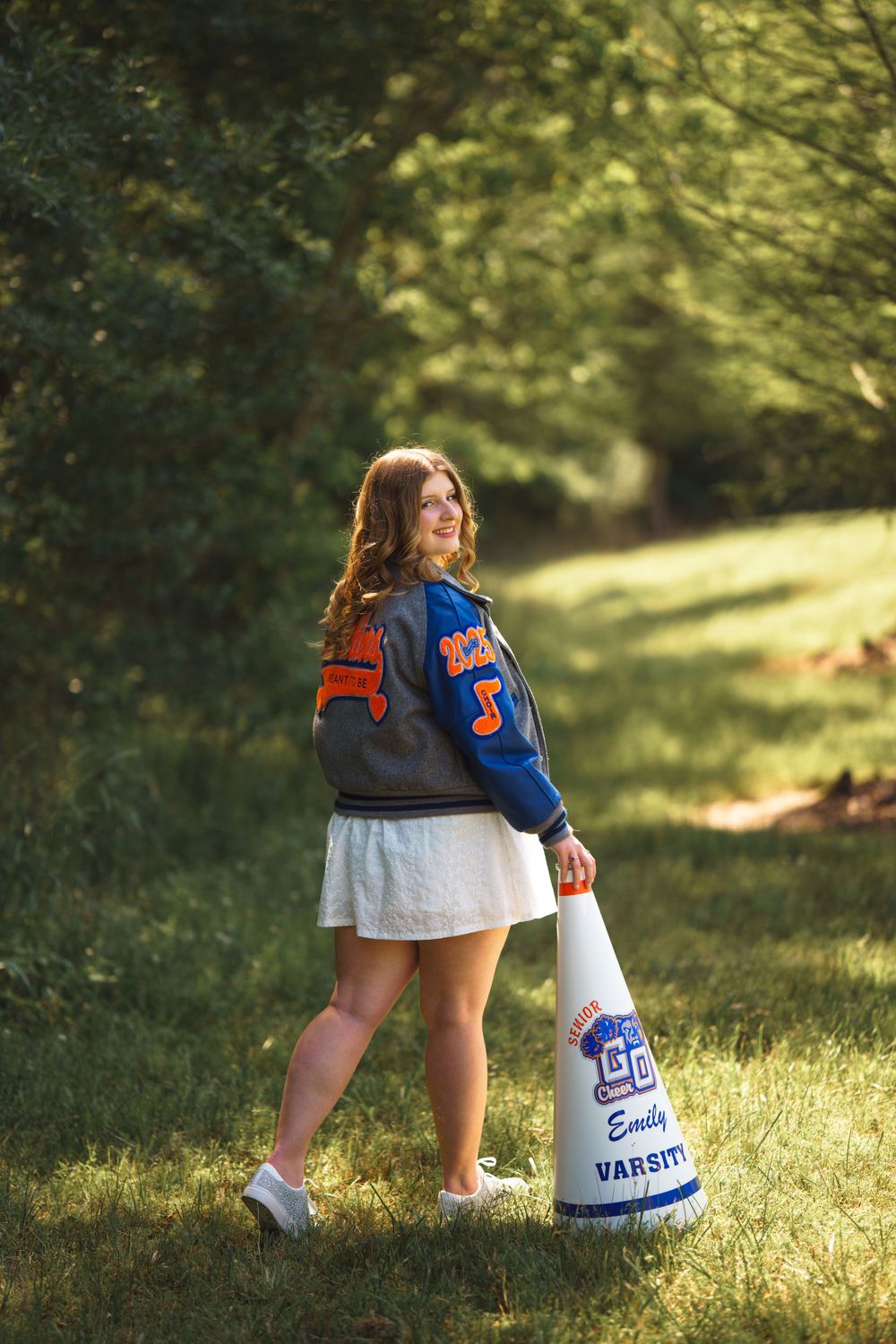 A student wearing a varsity letterman jacket stands in a sunlit grassy area holding a cheerleading megaphone.