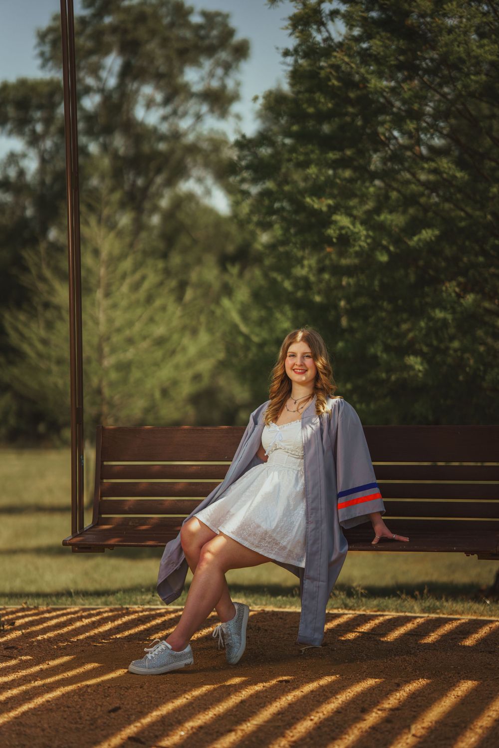 Person in white dress and varsity jacket posing on wooden bench in park setting with dappled sunlight.