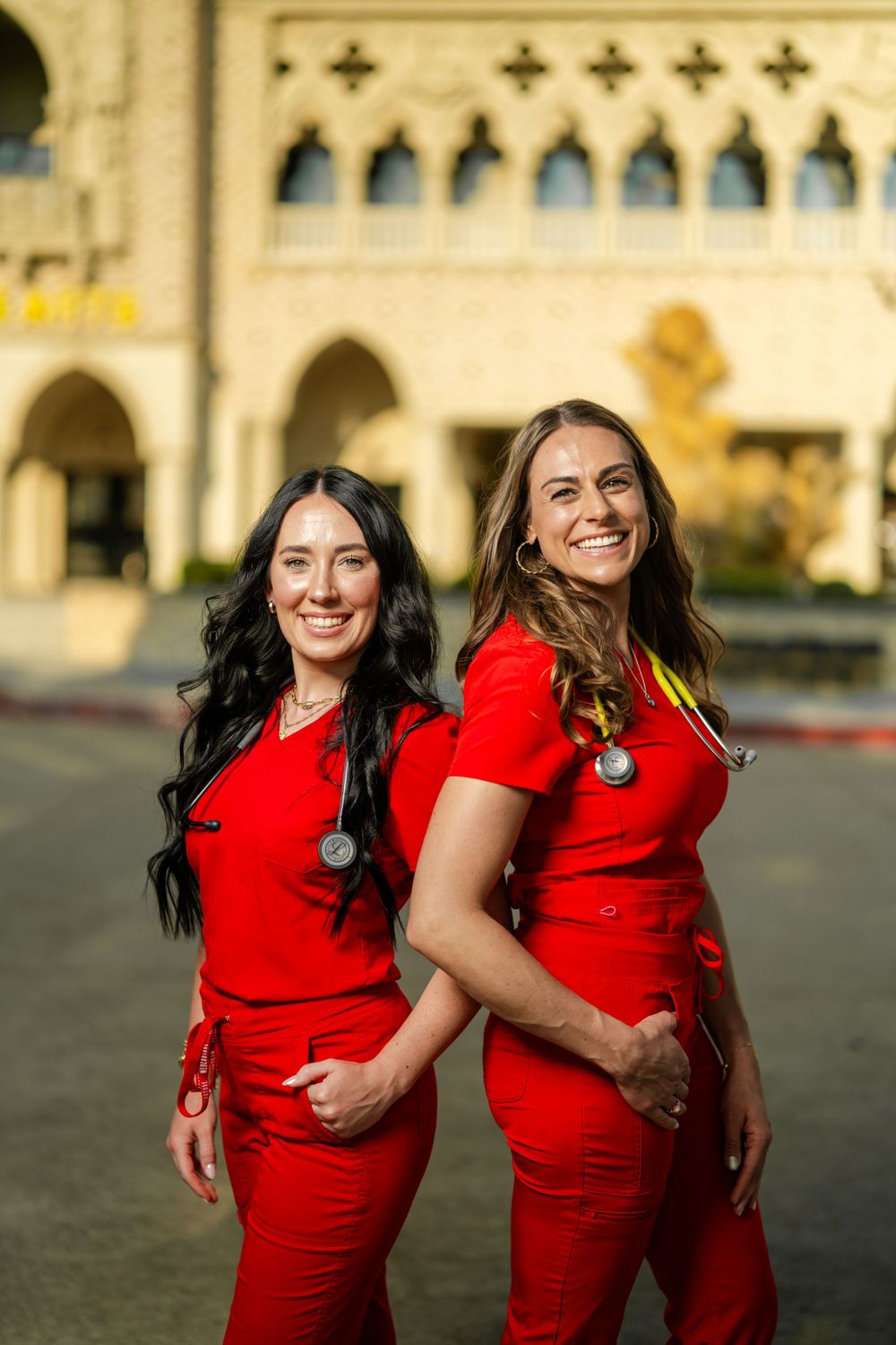 Two healthcare workers in red scrubs smiling while standing together outside a building with Gothic architecture.