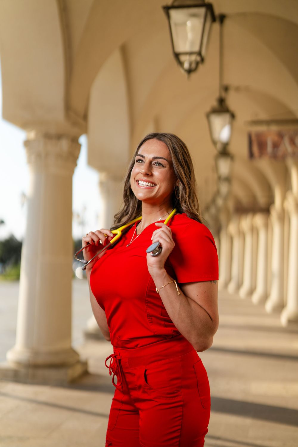 Woman in vibrant red dress poses elegantly under an arched colonnade with hanging lanterns at sunset.