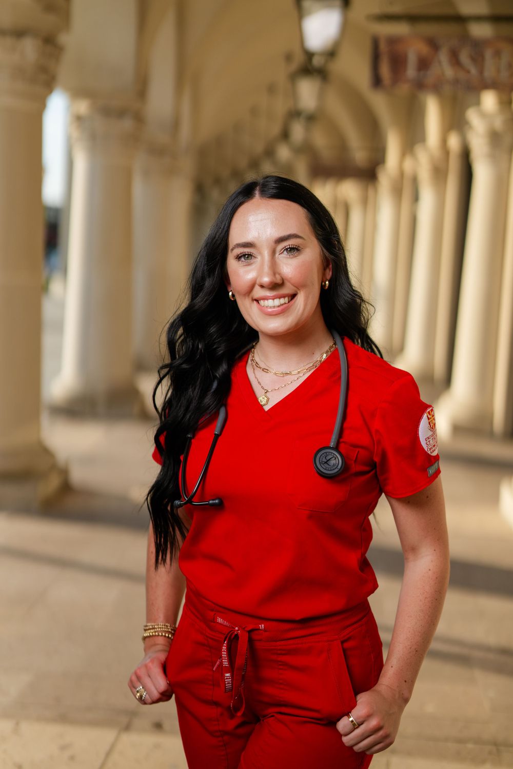 Healthcare worker in red scrubs with a stethoscope poses confidently in an arched corridor with classical architecture.