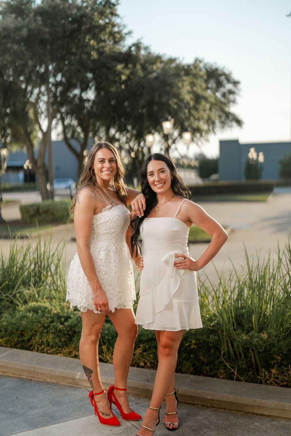 Two women in white dresses pose outdoors at sunset, one wearing red heels and the other wearing strappy sandals.