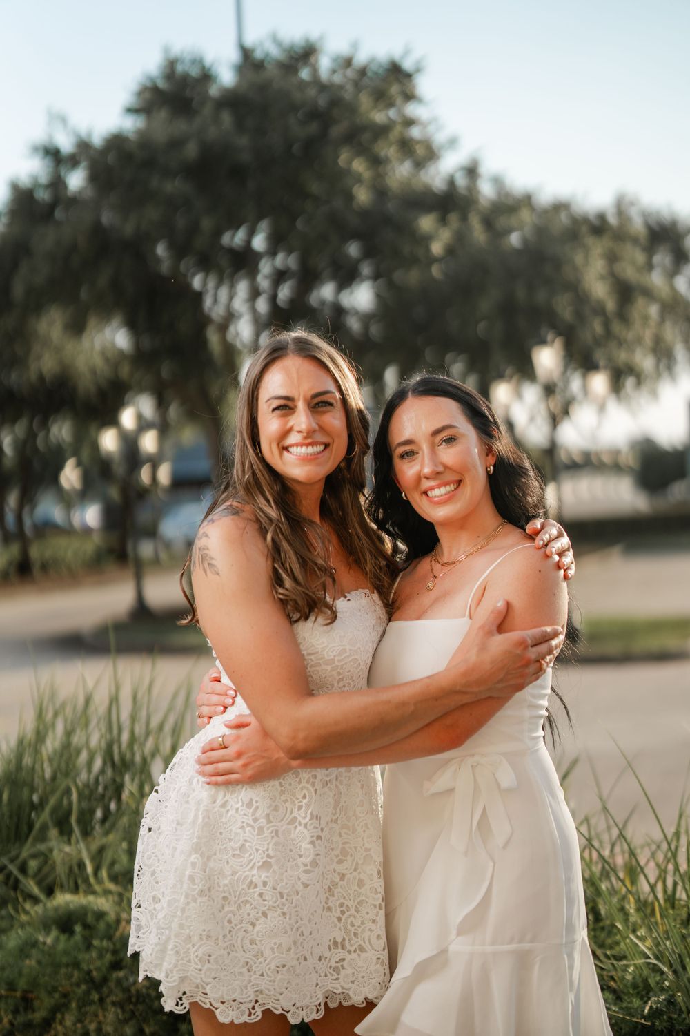 Two women embrace while wearing white dresses outdoors during golden hour with trees in the background.