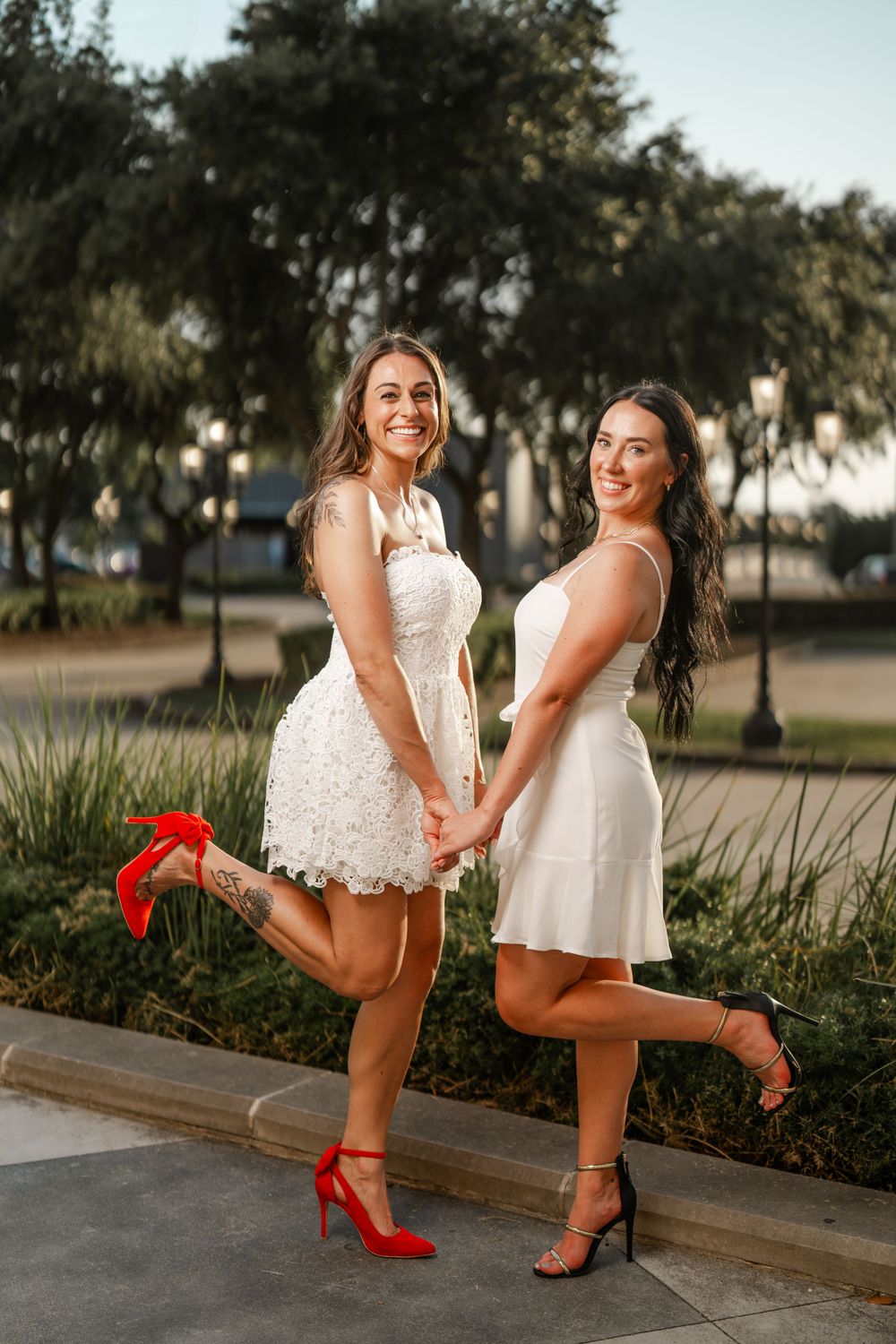 Two women in white dresses striking playful poses on a sidewalk at sunset while showing off their stylish heels.