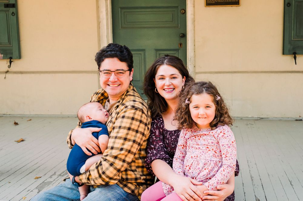 A family in casual attire sits together and smiles while posing on a white porch with green doors behind them.