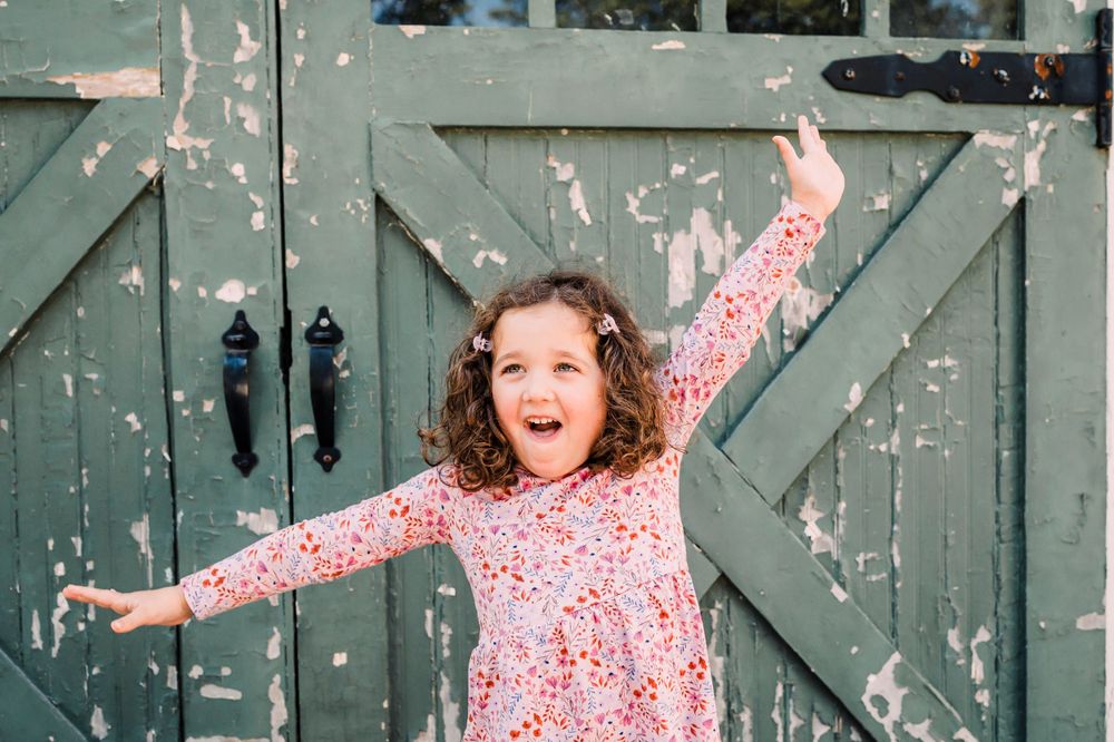 A young child wearing a pink floral dress jumps joyfully in front of a weathered green barn door.