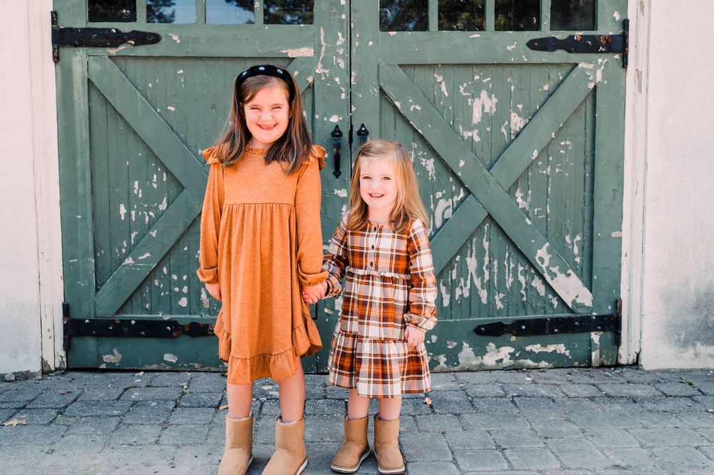 Two children standing together wearing fall-colored dresses in front of vintage green barn doors.