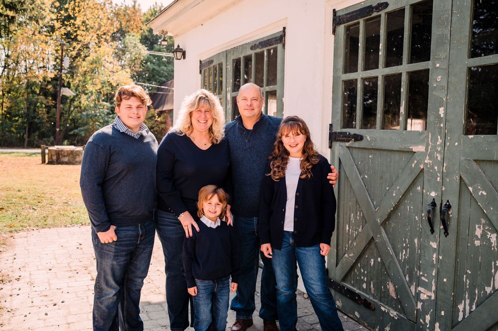 A group stands together smiling outside a rustic building with weathered barn doors on an autumn day.