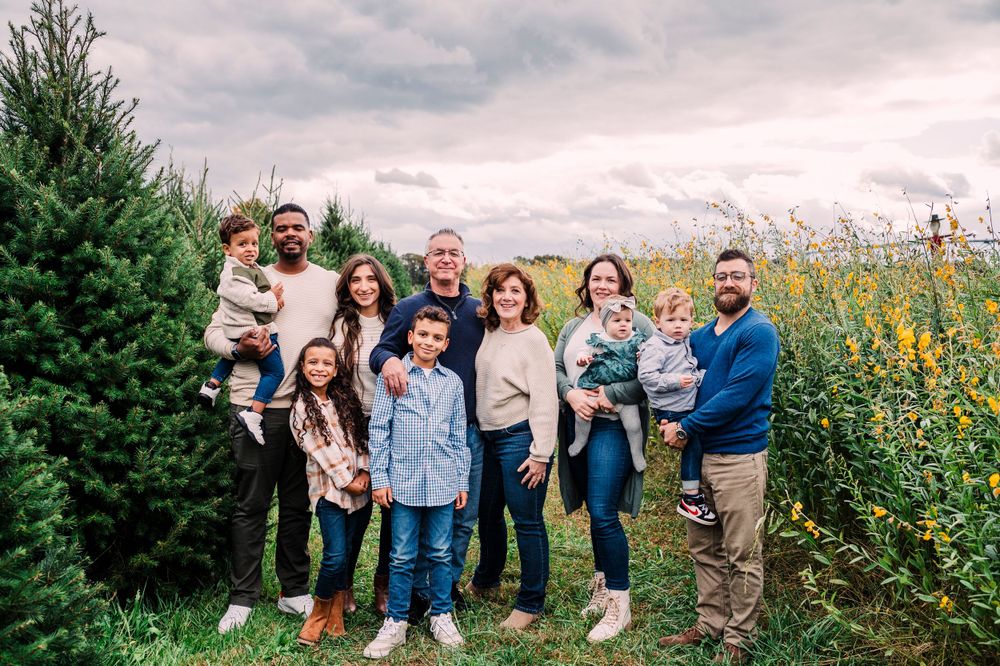 Family group standing together outdoors between evergreen trees and yellow flowers on a cloudy day.