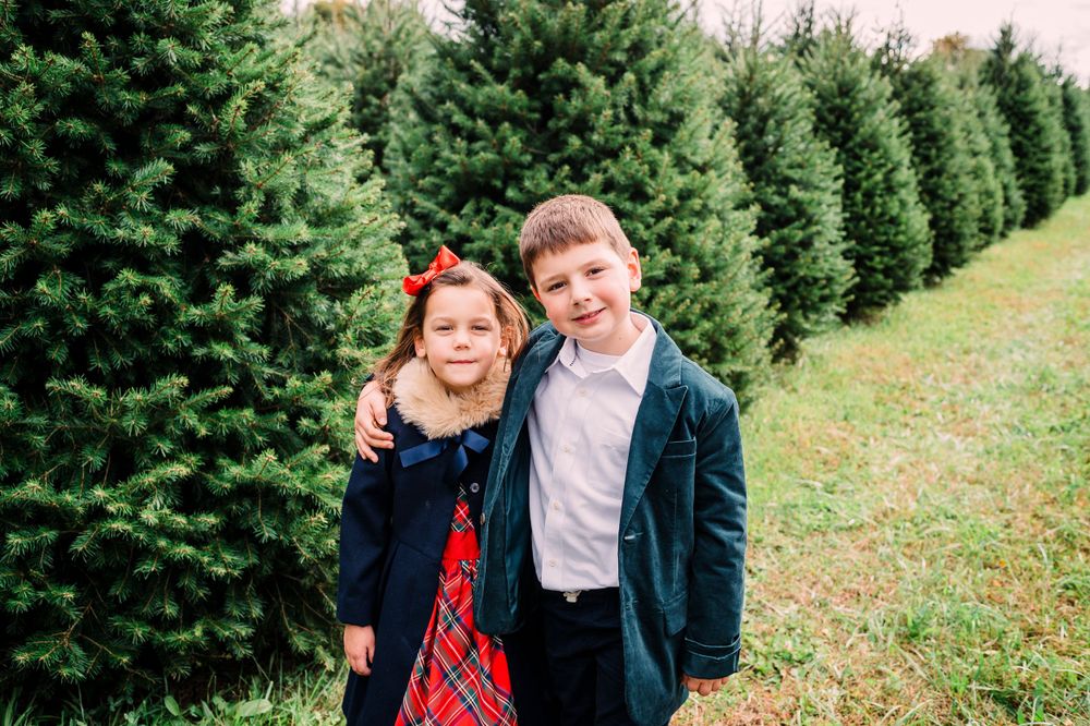 Two children standing together in front of rows of evergreen trees at a Christmas tree farm.
