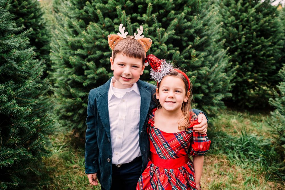 Two children wearing festive holiday outfits pose together at a Christmas tree farm with evergreens in the background.