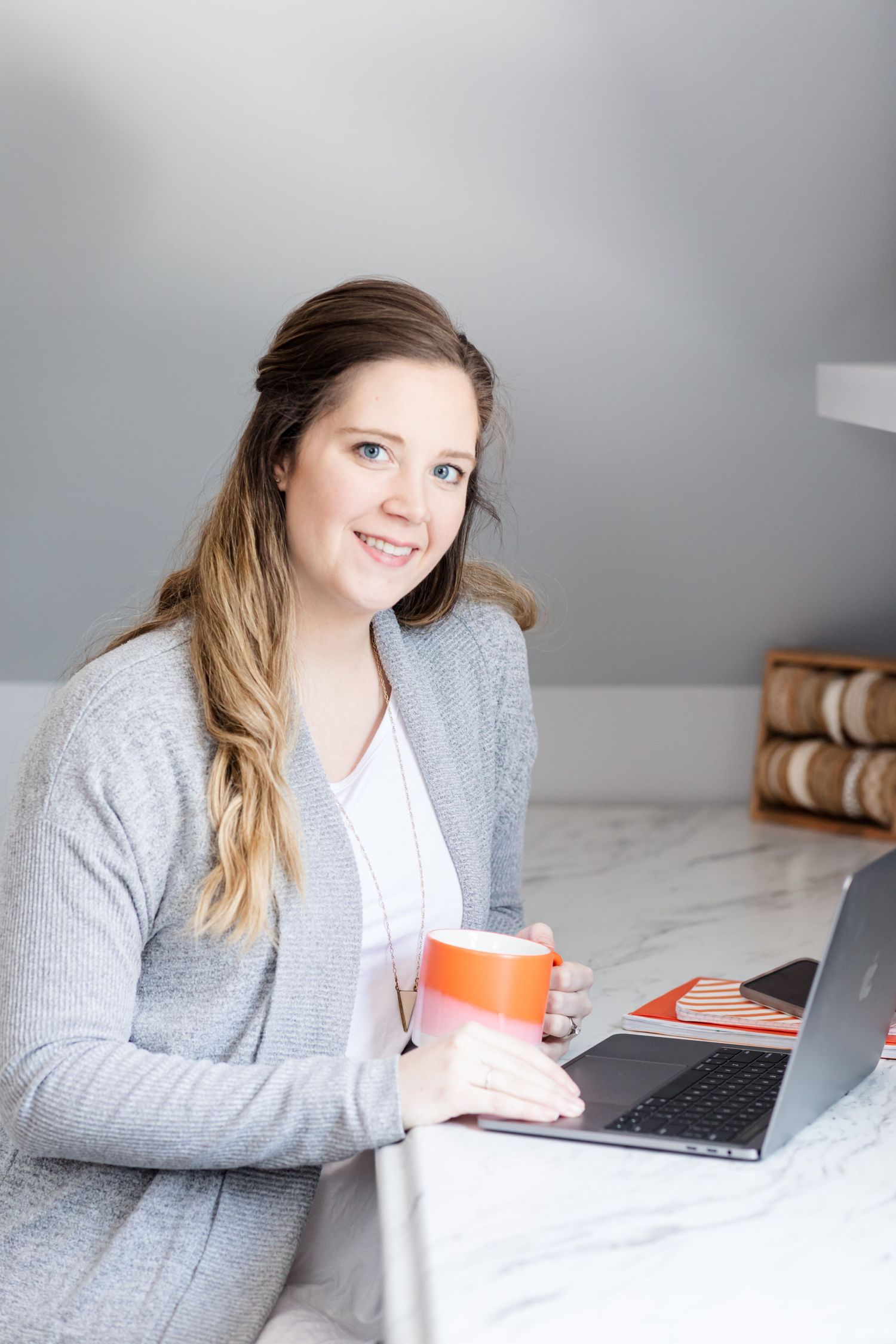 Person working on laptop with orange mug in cozy grey sweater against neutral background.