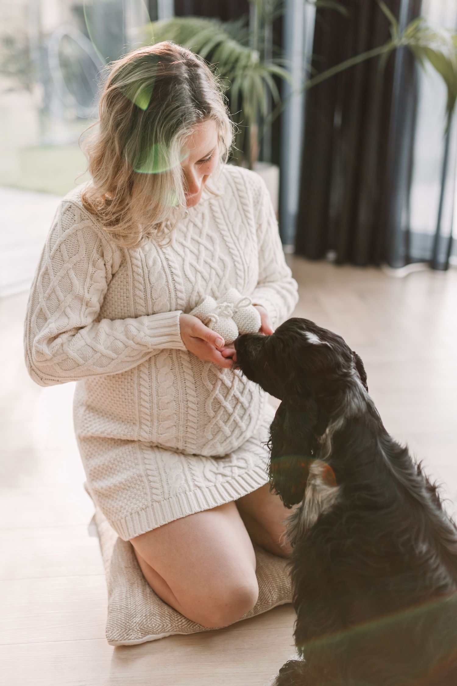 Pregnant woman in cream knit sweater sits on floor petting black dog in bright interior space.