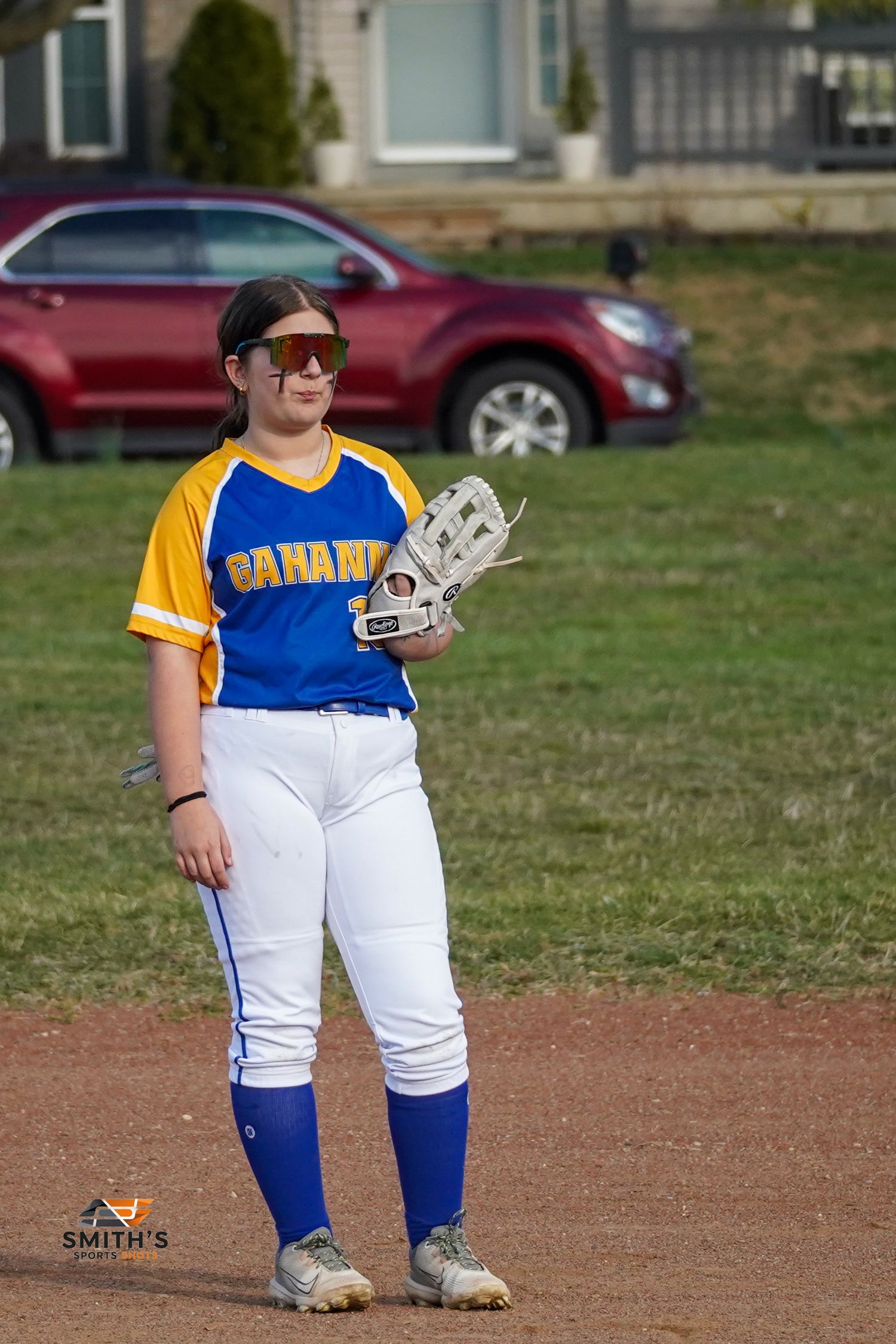 A softball player in a blue and yellow uniform stands ready with their glove on a dirt field.