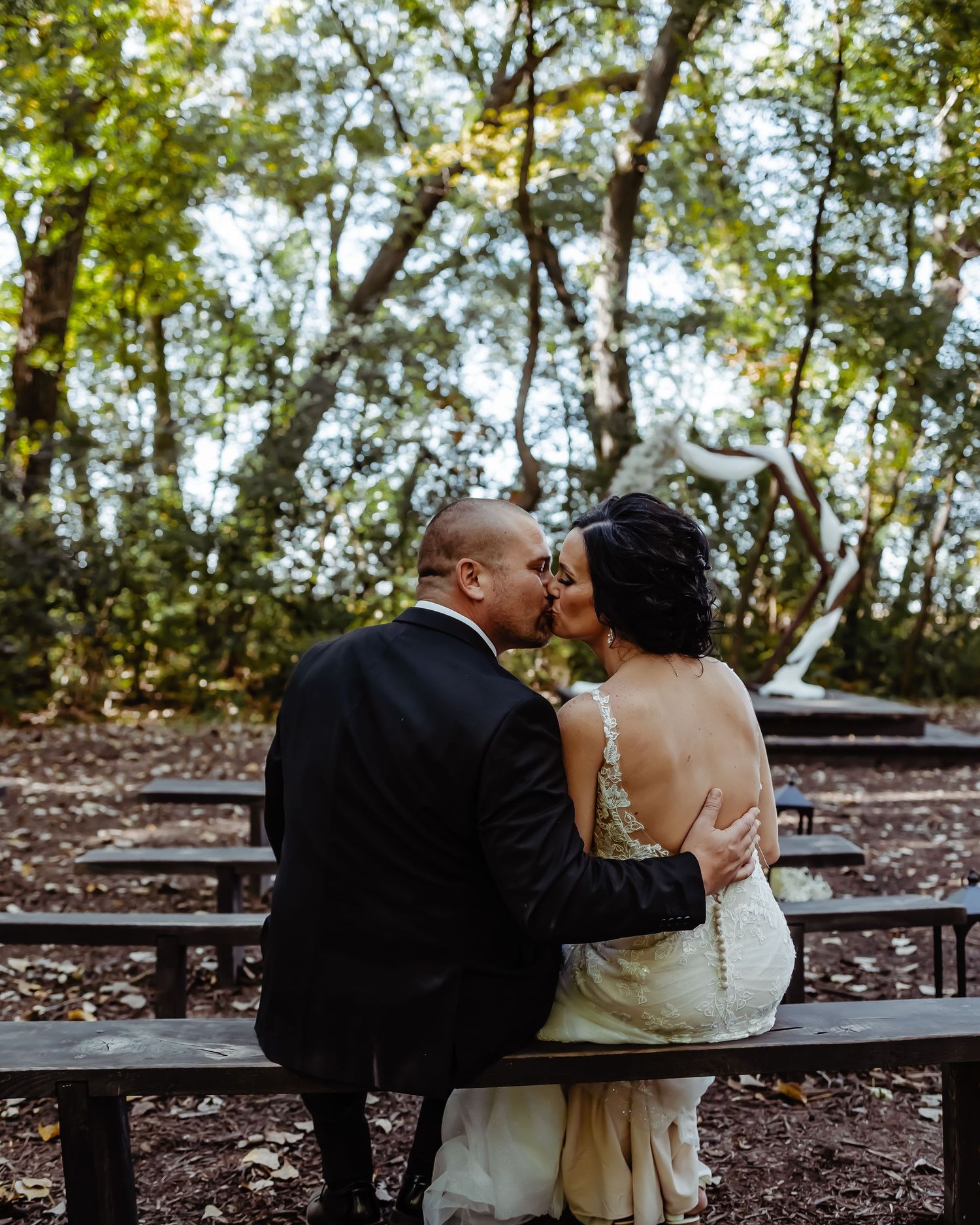 A couple shares a tender moment sitting on a bench along a wooded path during their wedding day.