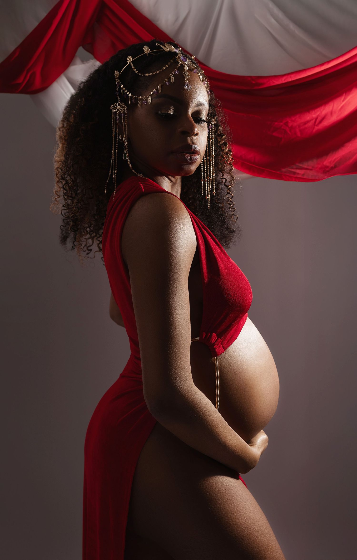 Artistic maternity portrait with flowing red fabric and dramatic lighting showcases an elegant silhouette against gray backdrop.