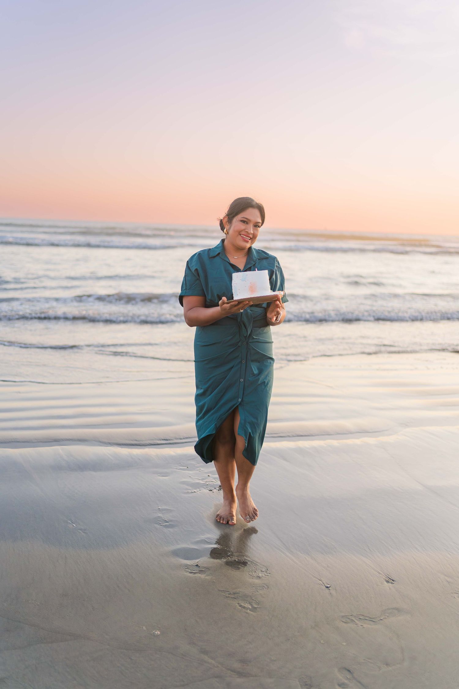 Person in teal dress walks along the wet sand at sunset while ocean waves roll in.
