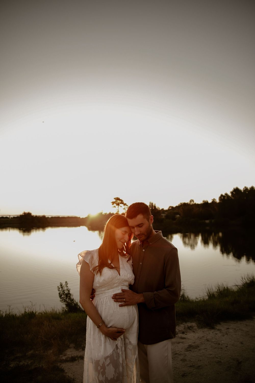 Sunset maternity photo by lake shows couple in tender embrace.