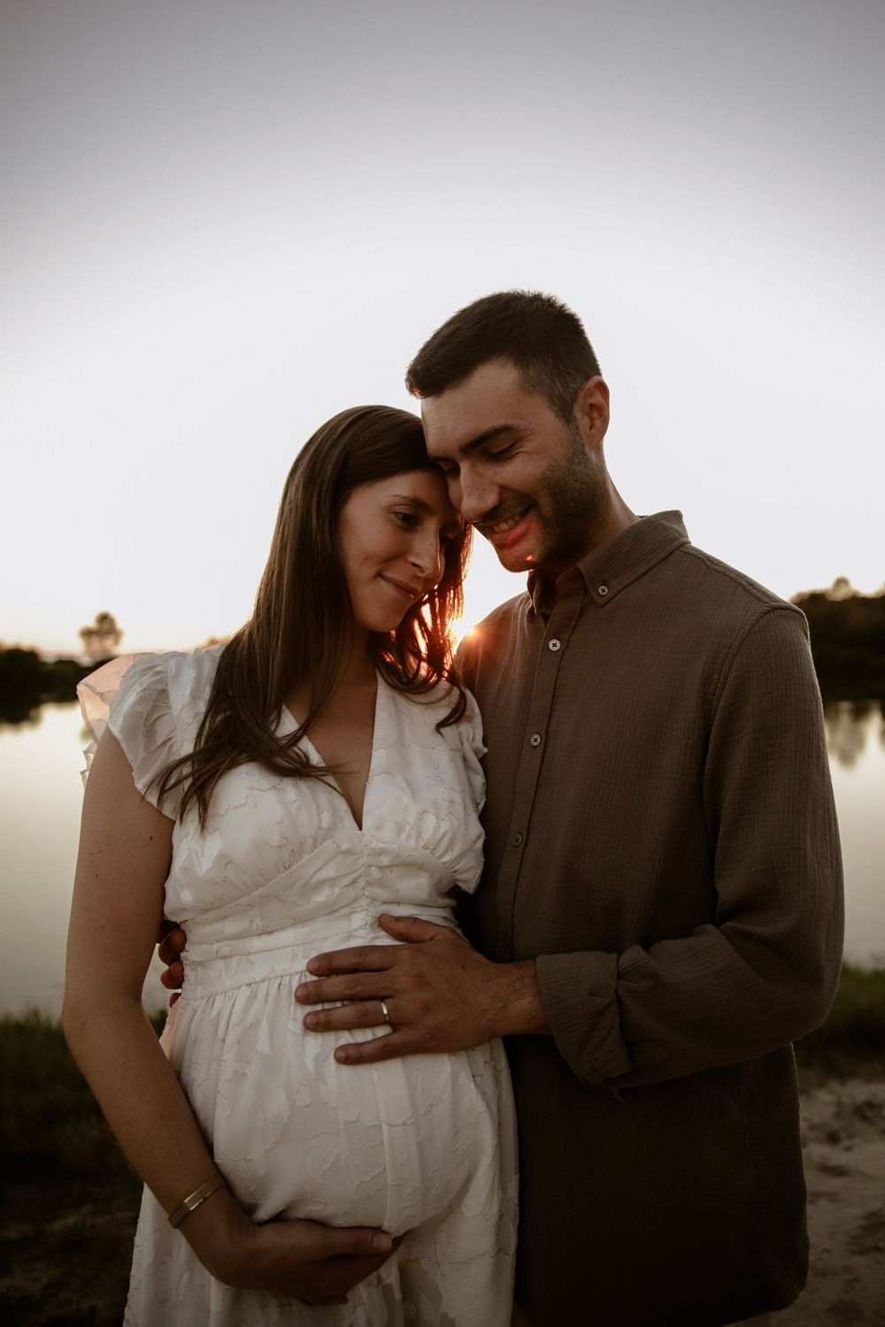 An expectant couple embraces tenderly during their maternity photoshoot at golden hour near water.