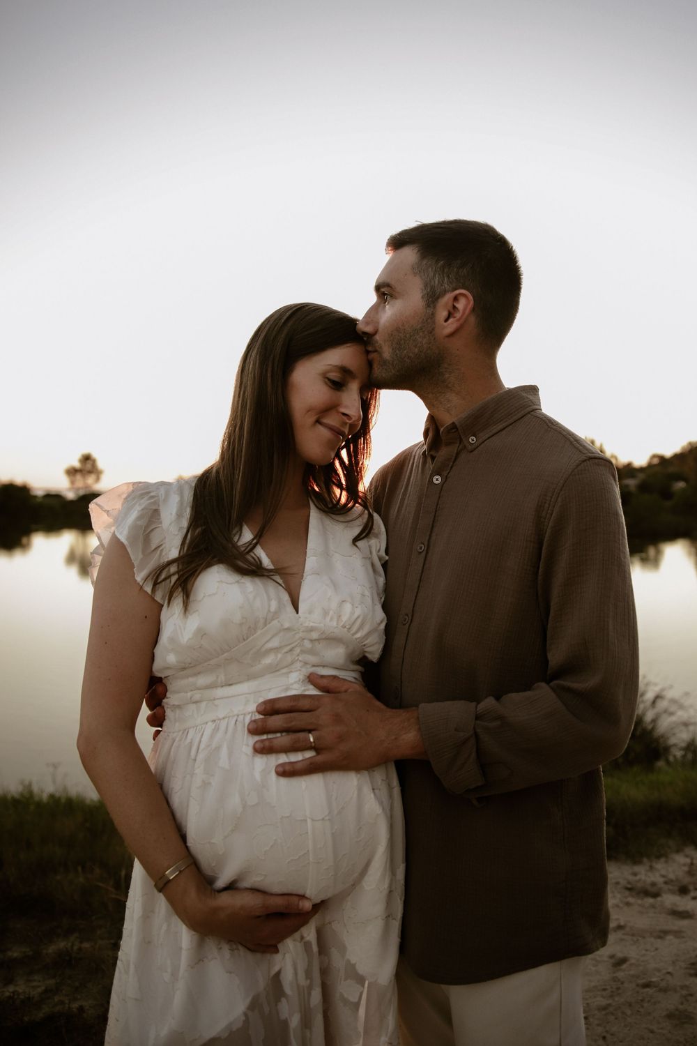Couple shares intimate moment during waterfront maternity photoshoot at dusk.
