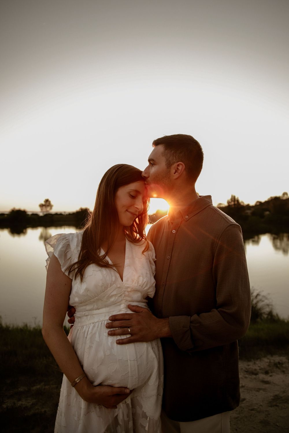 Couple embraces during sunset maternity session by lake with golden sunlight behind them.