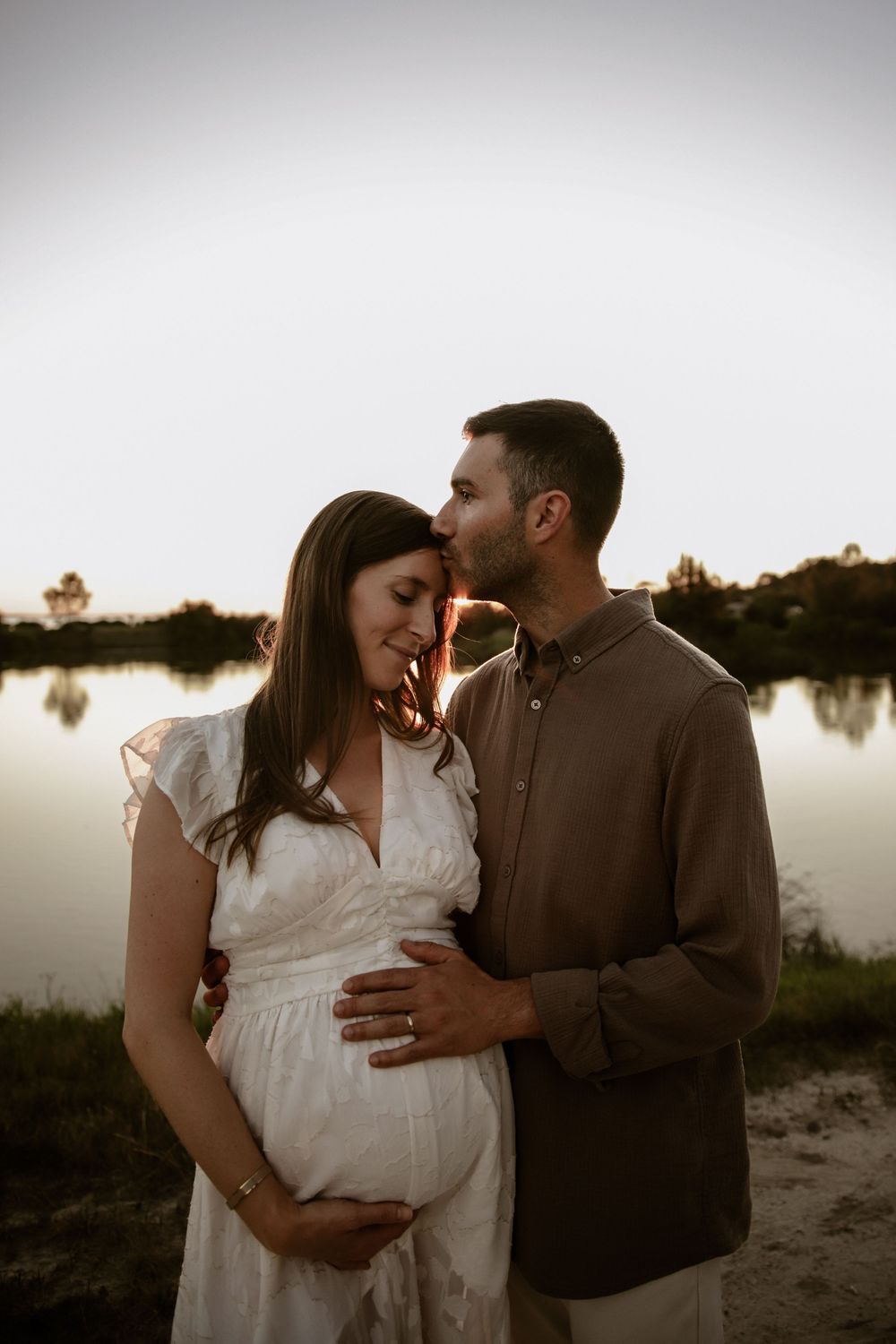 A romantic maternity portrait captures a tender moment between a couple at dusk by a lake.