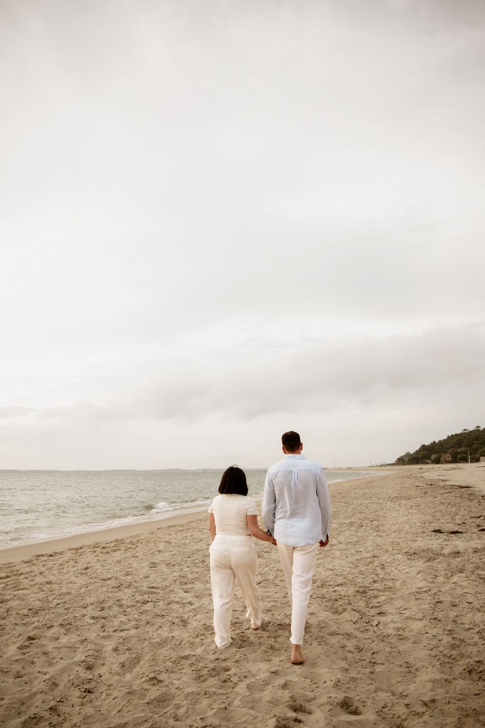 Couple in white outfits walking together along a serene beach shoreline on an overcast day.