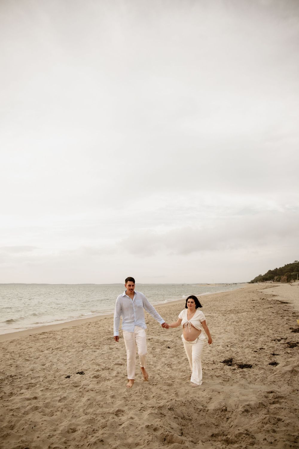 A couple in white outfits holds hands while walking along a misty beach shoreline in a romantic scene.