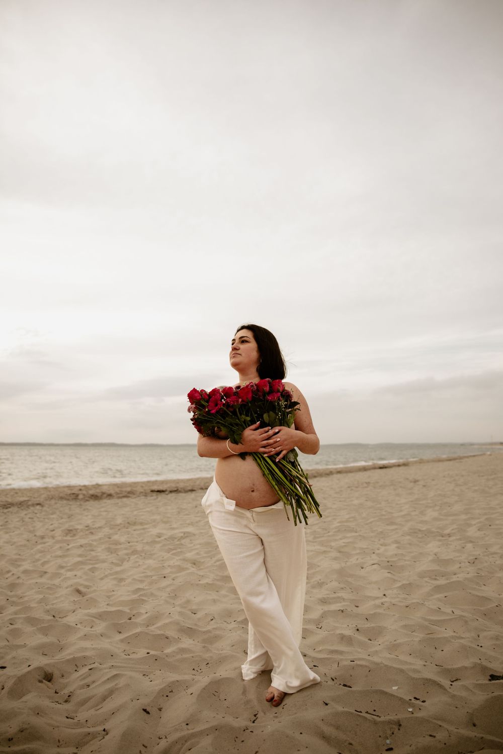 Woman in white dress holding red roses strikes dramatic poses on a cloudy beach.