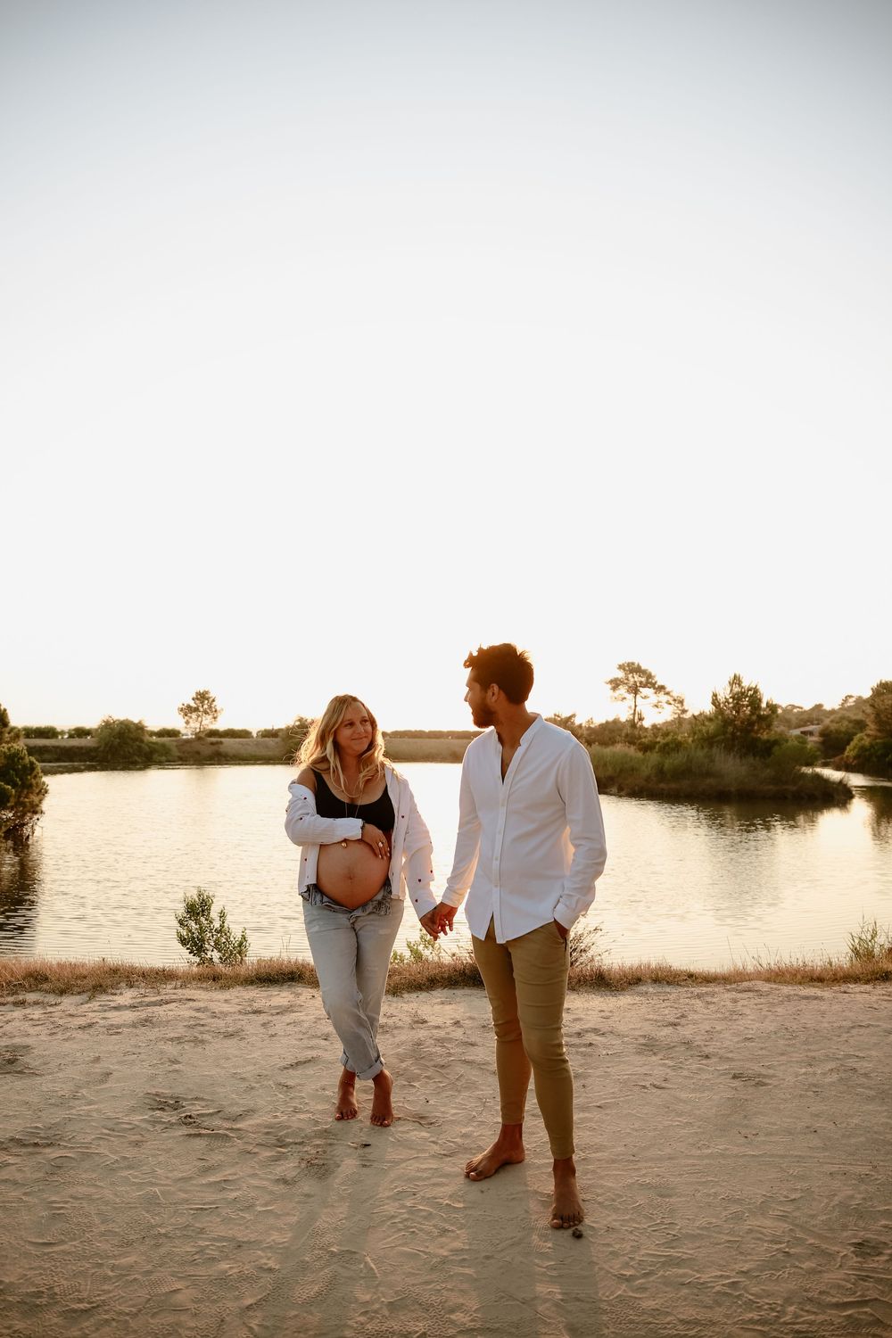 Couple holds hands during sunset photoshoot at sandy beach near tranquil lake.