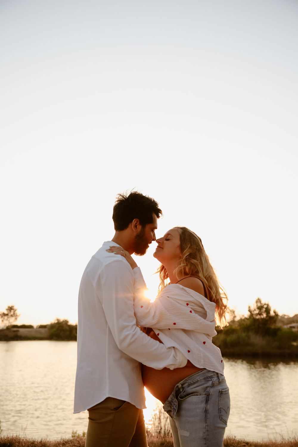 A romantic couple embraces by a lake at sunset with golden light illuminating their silhouettes.