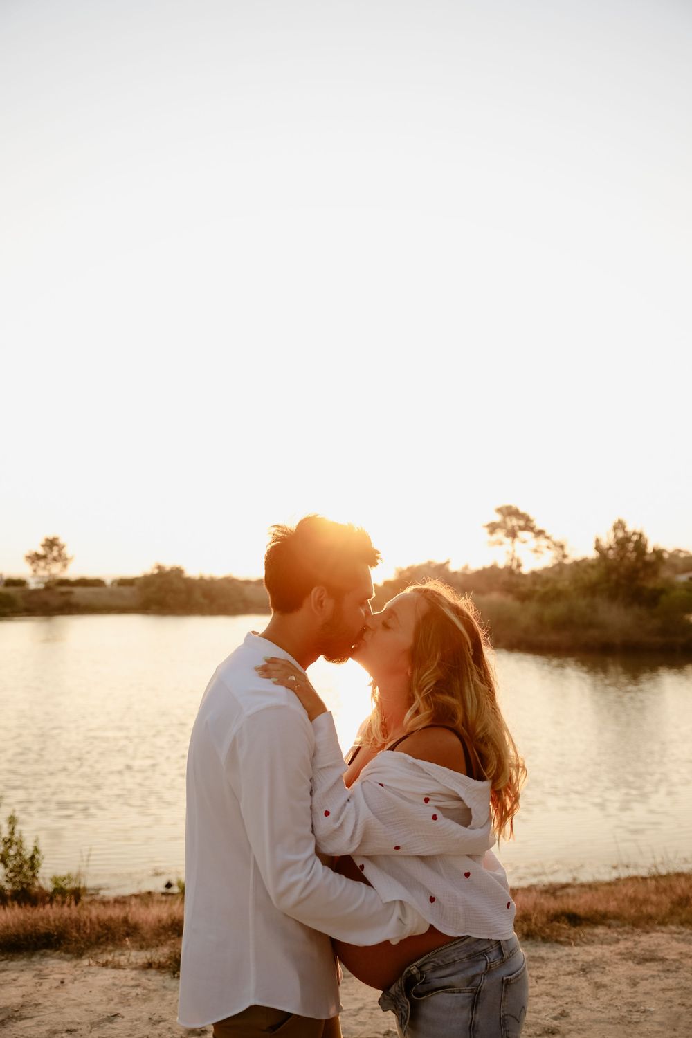 A romantic couple embraces on a beach at sunset with a lake and trees in the background.