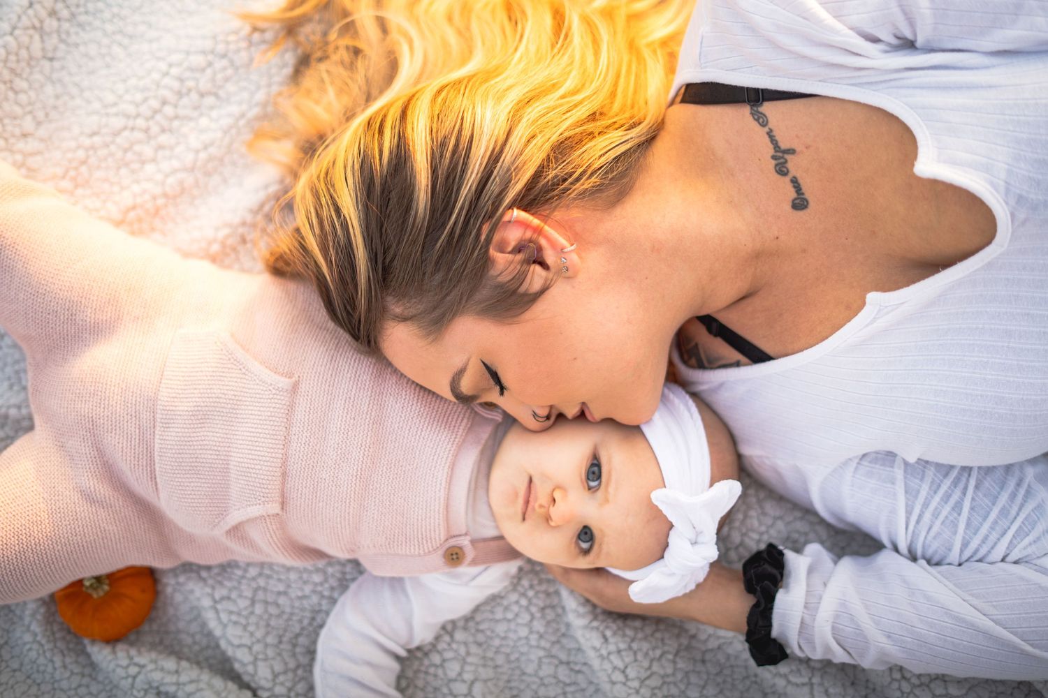 a mother laying on a blanket with her daughter. she looks behind to give her a kiss while she plays with her pumpkin