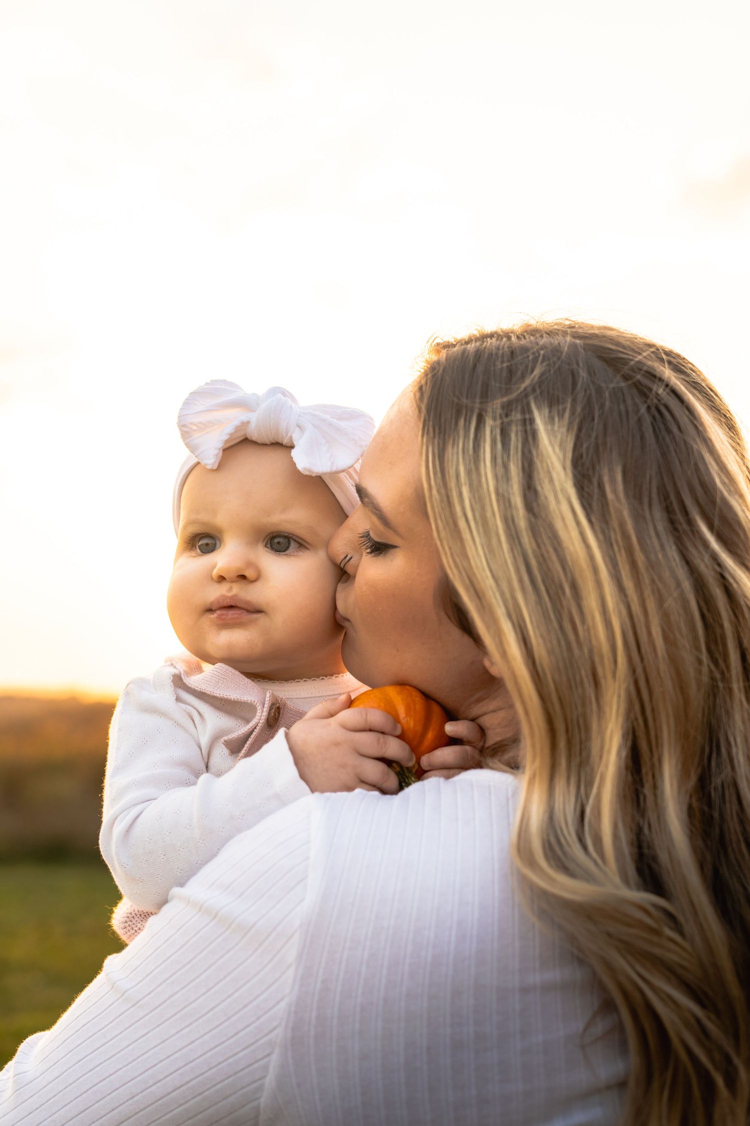 a mother kisses her daughter while holding her. shes holding a pumpkin. the sun is shining during a beautiful golden hour