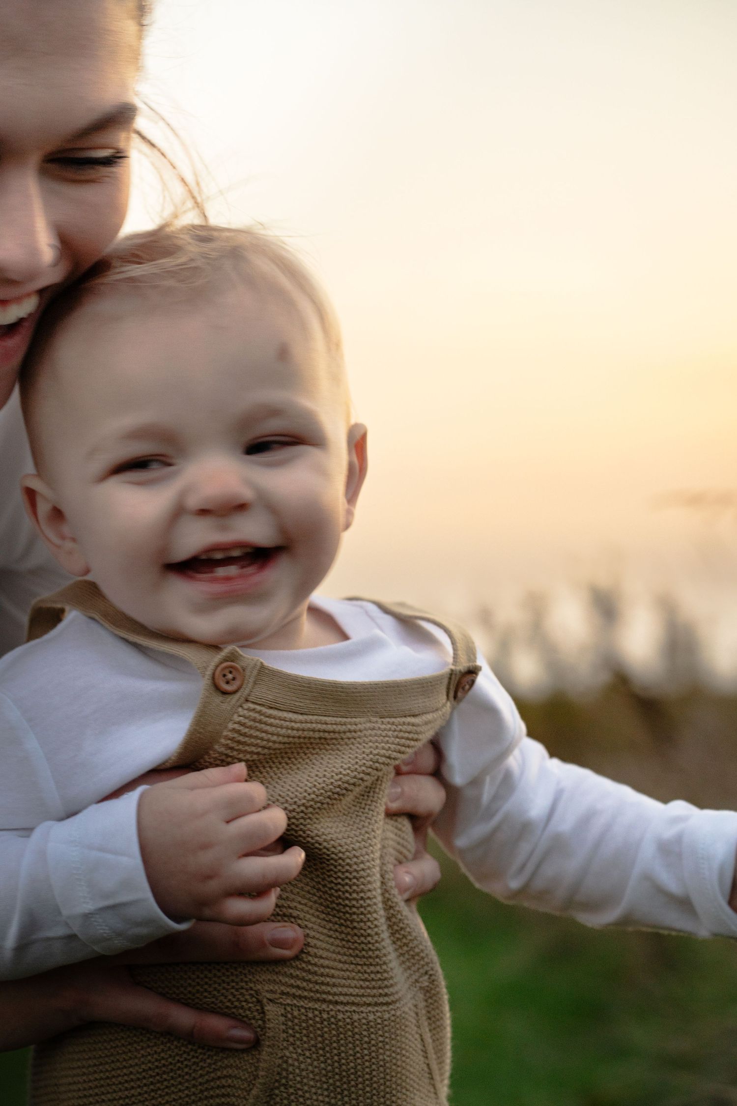 a baby laughing and smiling in a field while his mom lifts him up. sun is shining from behind with a warm, golden hue