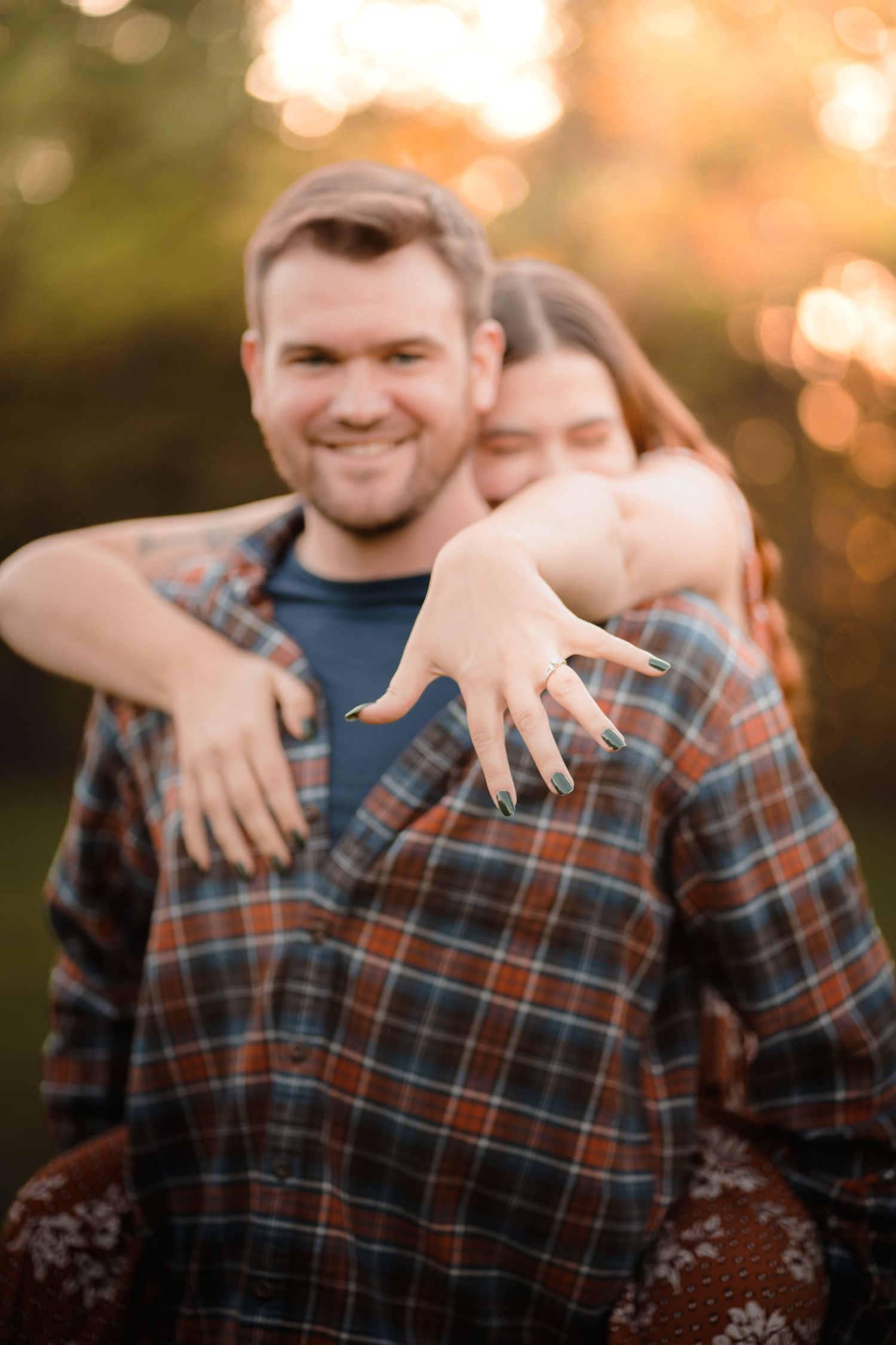 an excited new fiancee jumping on her new fiance's back while holding her hand out in front of them both into the camera to show her ring