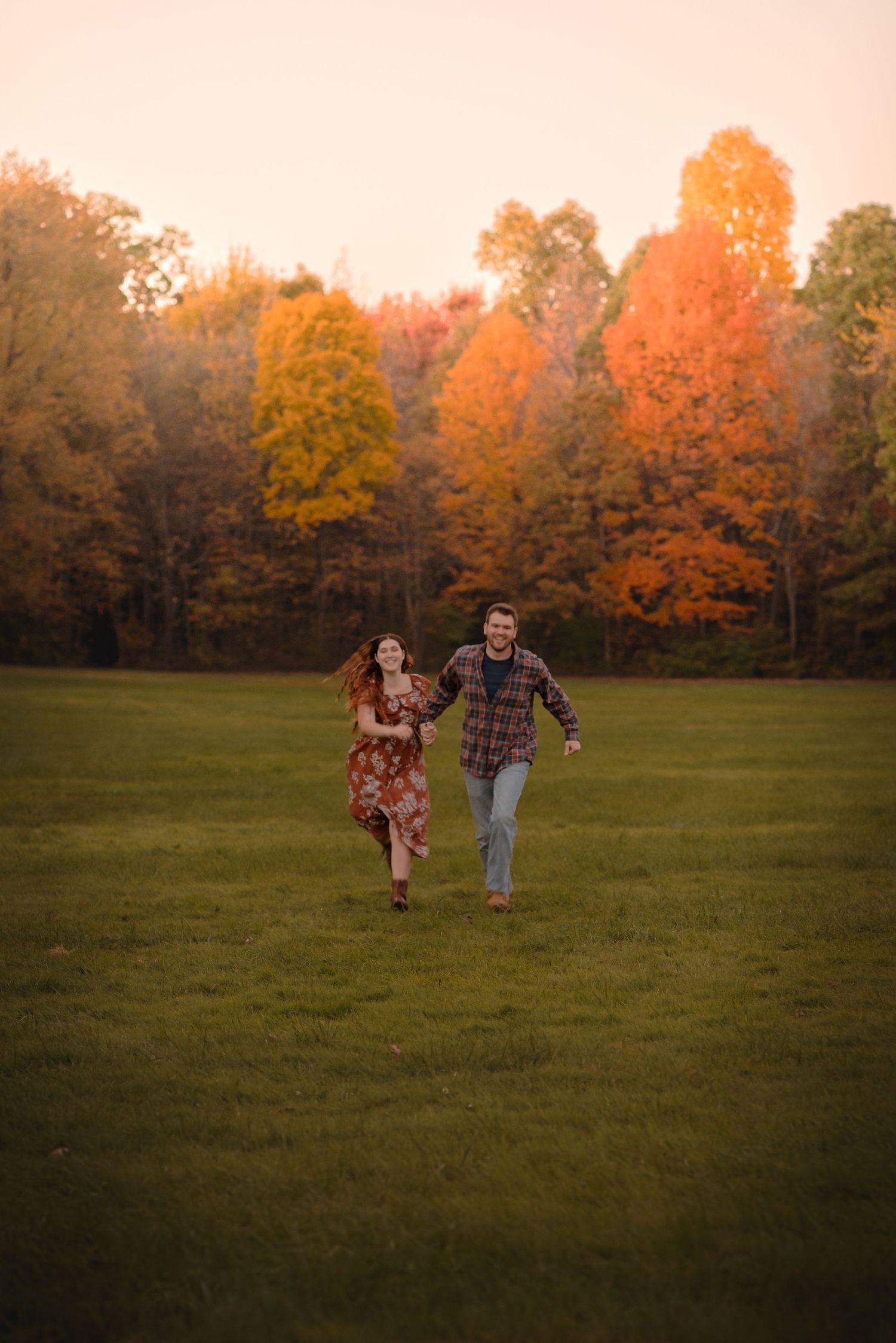 a newly engaged couple running through a field and towards the camera while holding hands. the trees behind them are very colorful