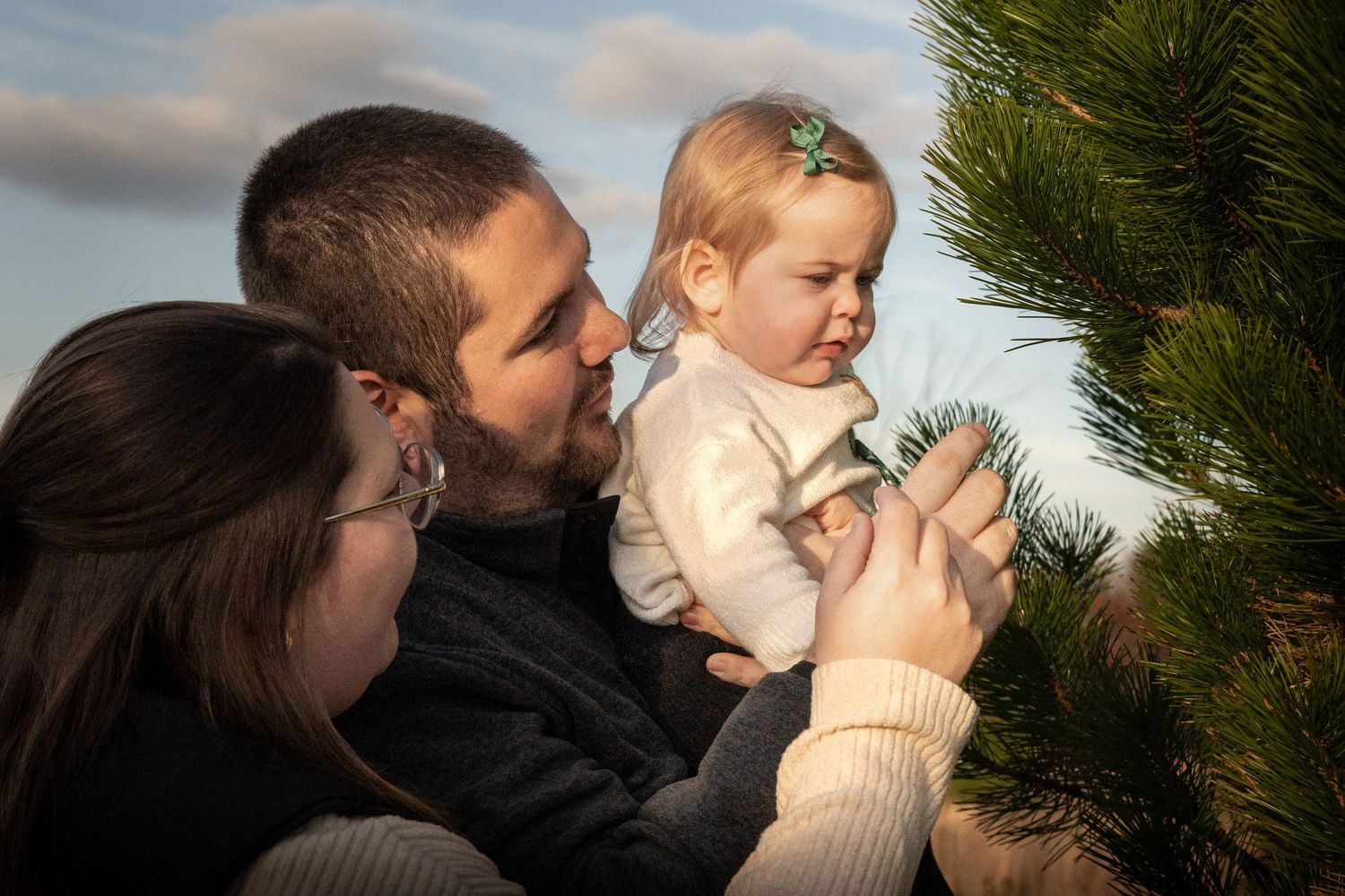 A couple shares an intimate kiss outdoors with a cloudy sky and evergreen trees in the background.