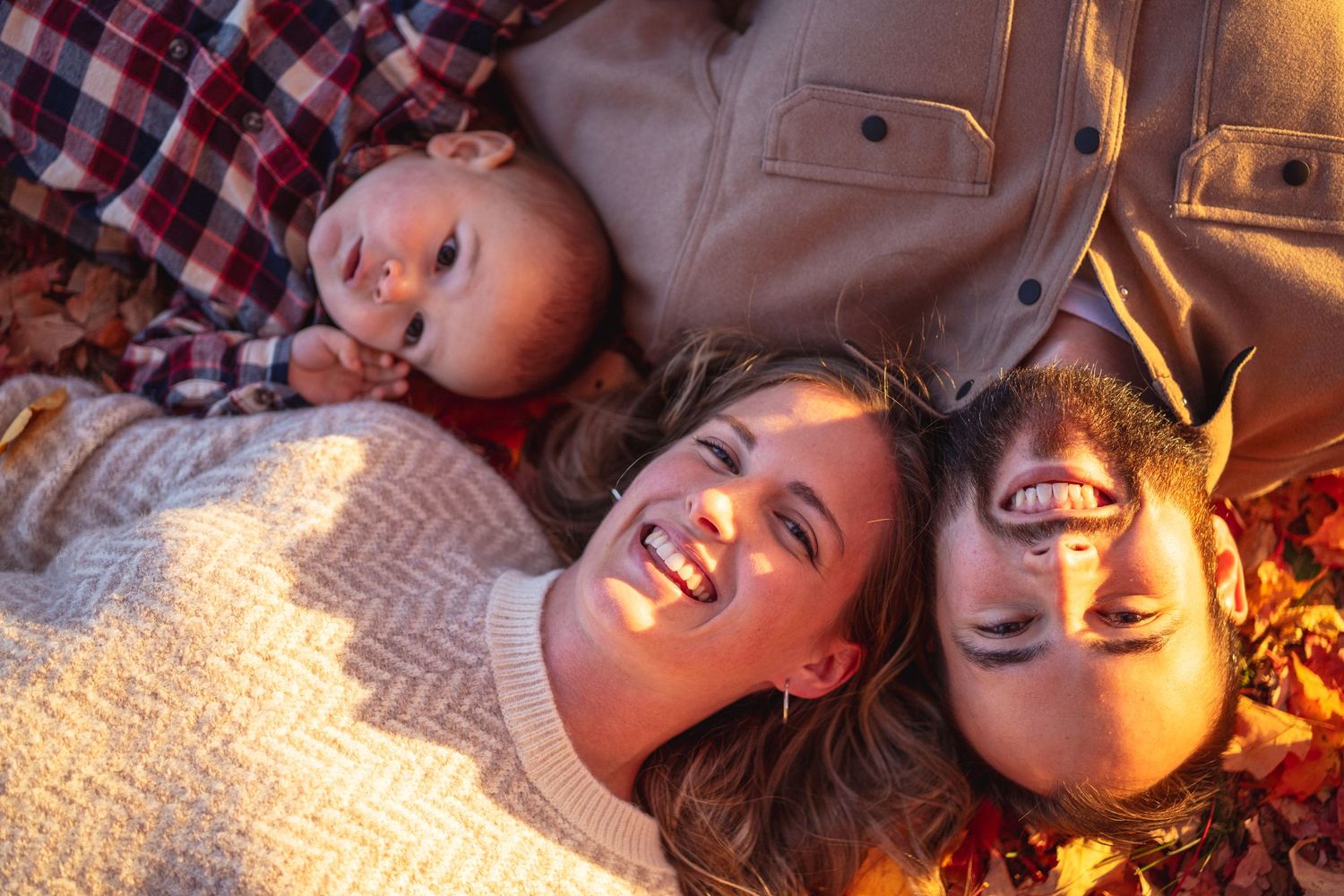a happy family laying on the leaves on the ground, looking up and smiling at the camera during golden hour for a fall family session