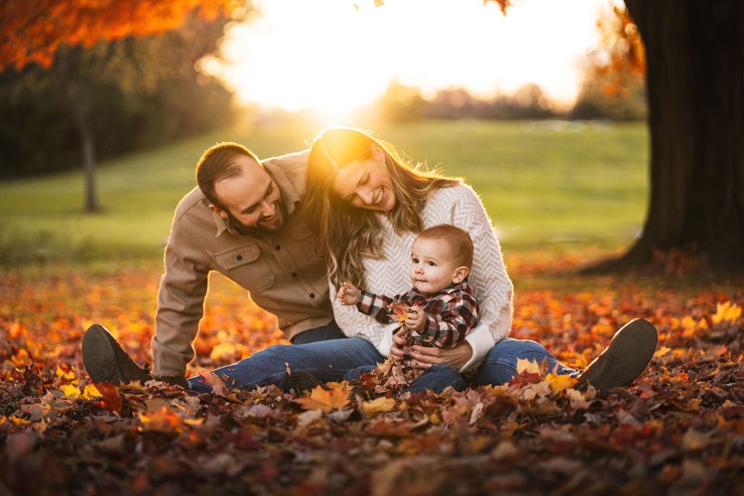 a young family and their toddler sitting and playing in the fall leaves. the sun setting under the horizon in the background