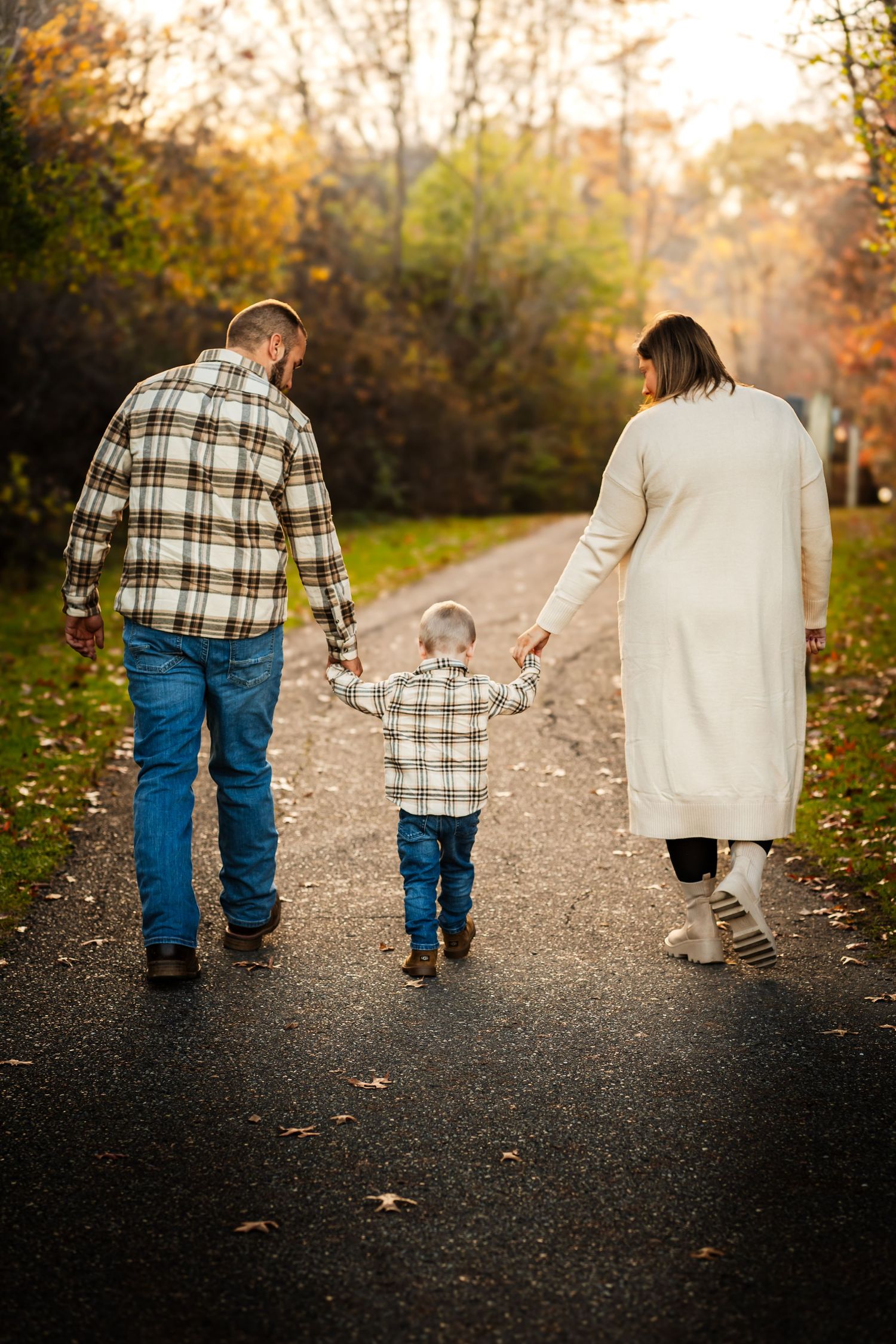 a family walking on a path surrounded by trees. both adults are on each side holding the sons hands, the trees are starting to change color