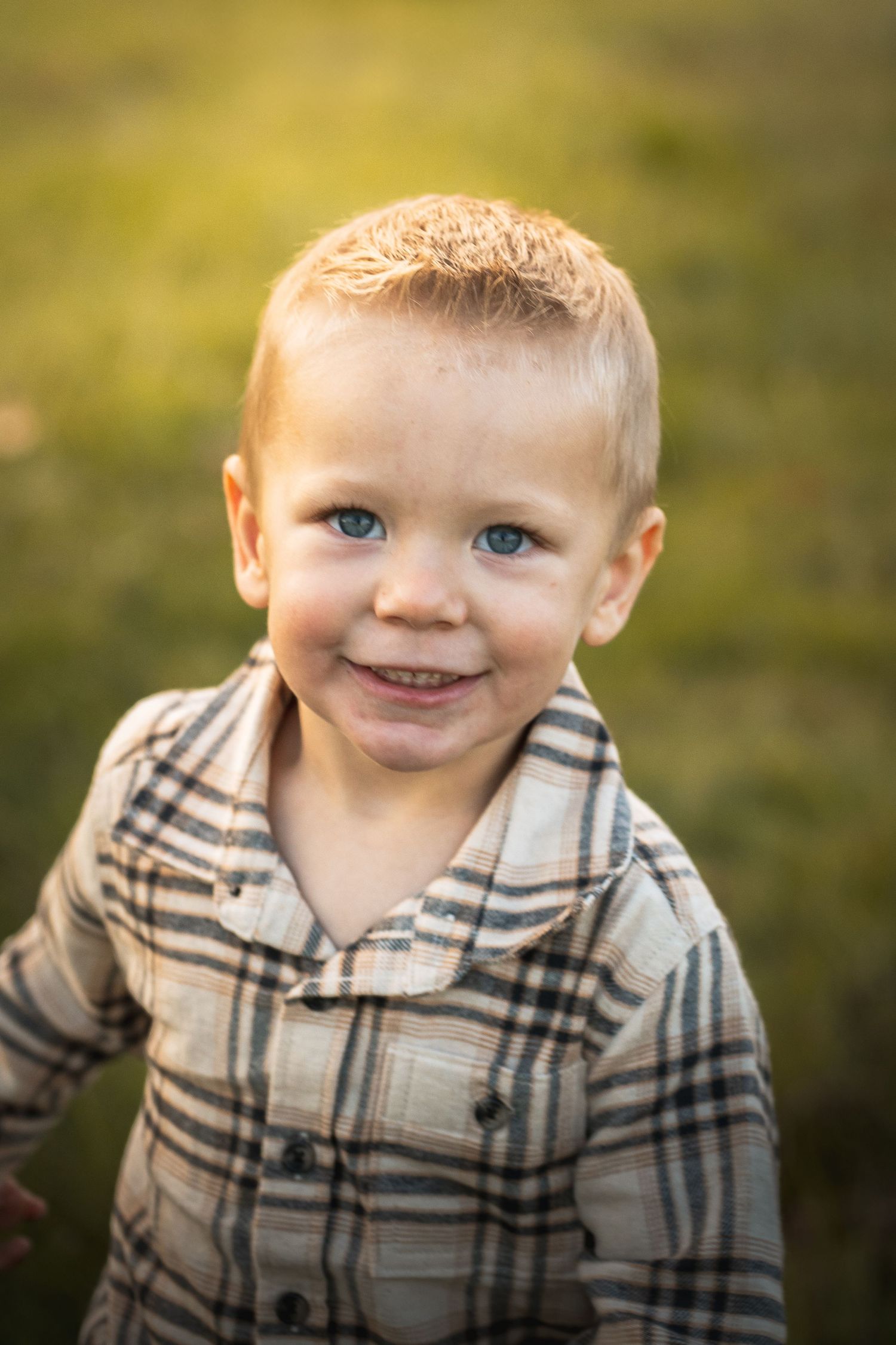 a boy looking up and smiling at the camera while running through a green grassy field. the sun shining off of the left side of his flannel
