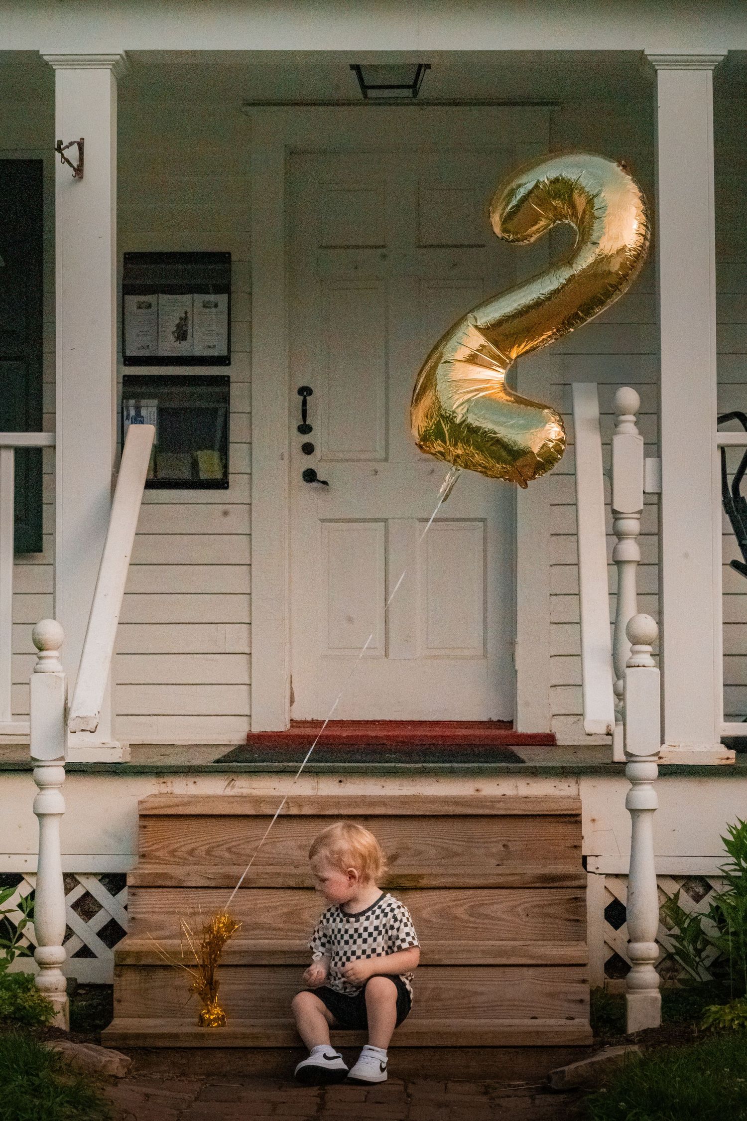 Someone sits on a wooden porch of a white house with gold number 2 balloons floating in the air.