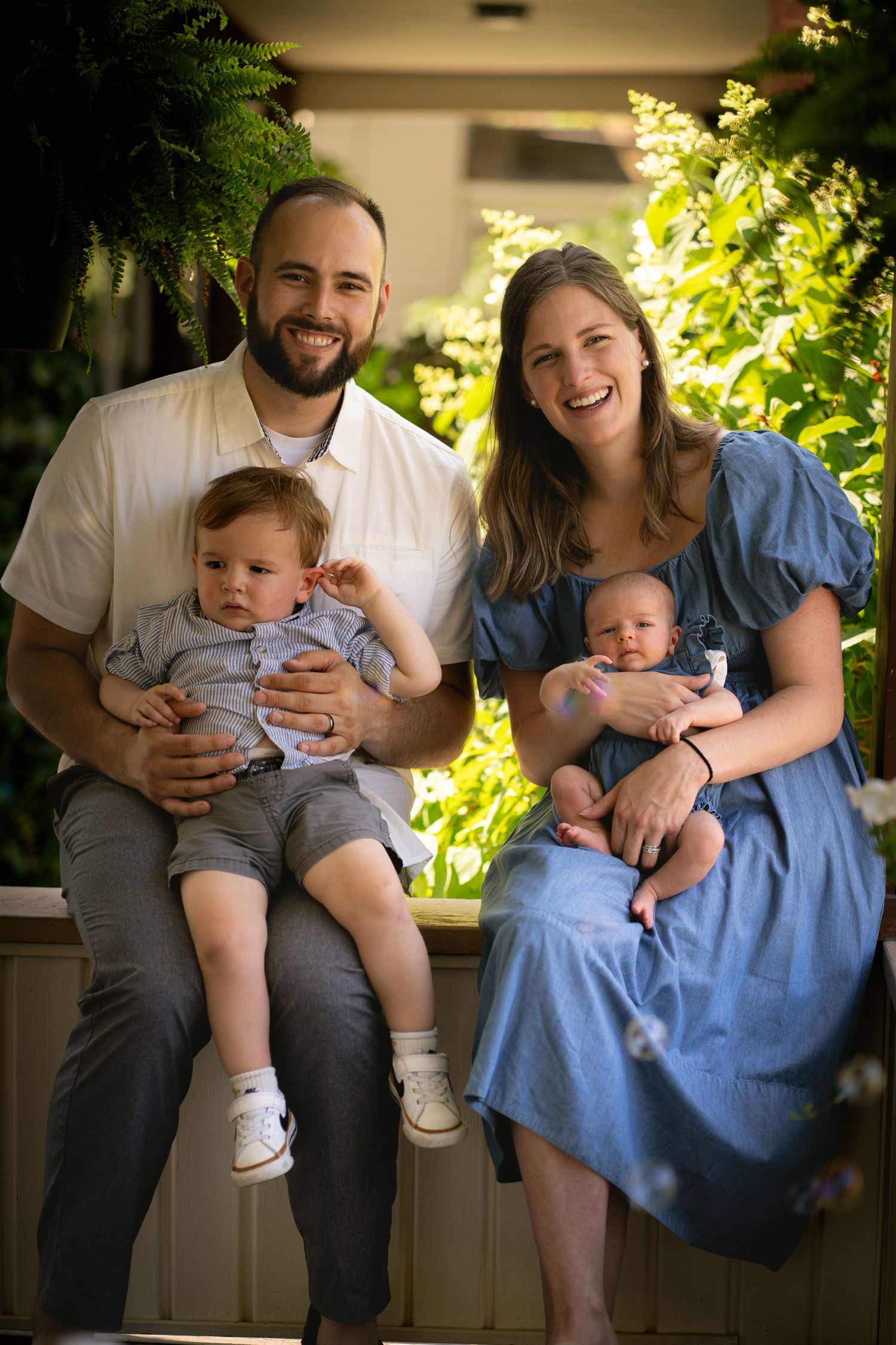 Young couple sits with infant in natural light beside window, sharing tender moments together in series of intimate portraits.