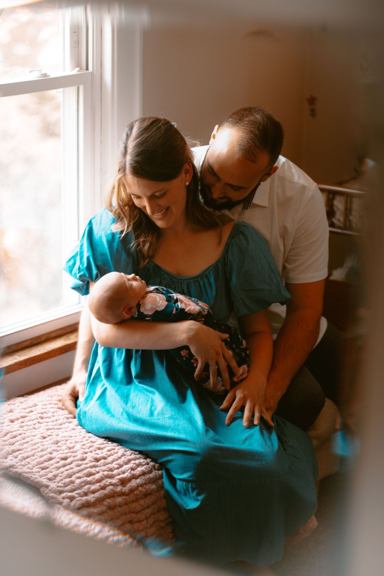 Parents share an intimate moment with their newborn baby while sitting by a window in a warm, sunlit room.